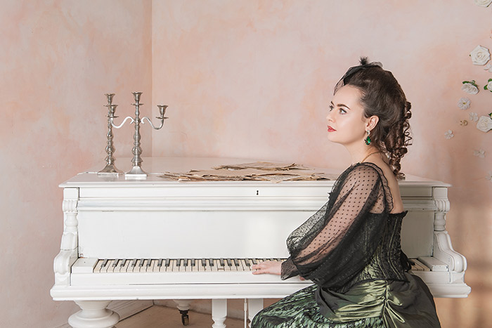 Beautiful woman wearing green victorian style dress sitting near piano feat